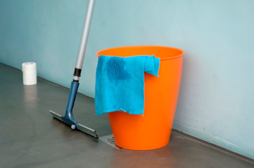 Close-up of cleaning supplies including orange bucket, mop, and blue rag indoors.